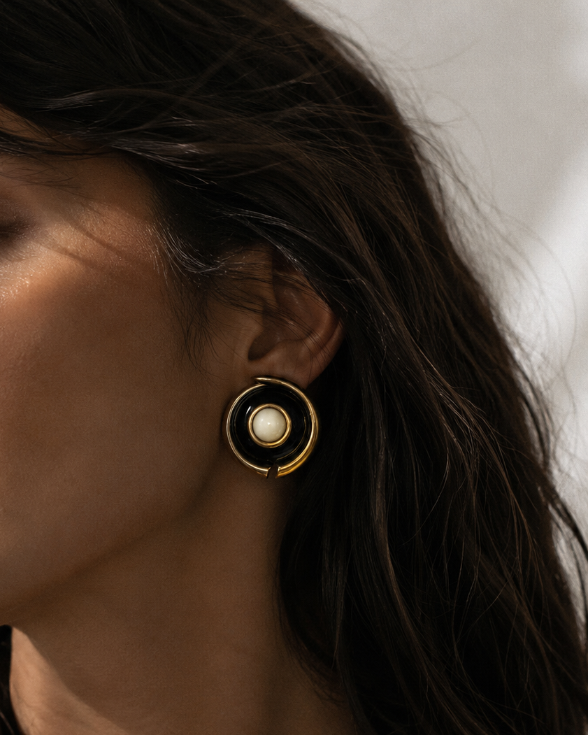 A close-up of a woman wearing a gold and black circular drop earring with a white pearl center, shown in profile against a soft light background.
